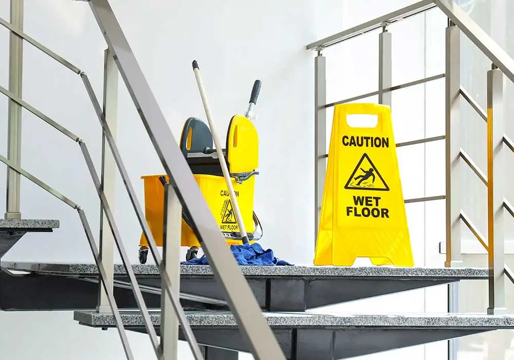Impresa di Pulizie Antonella Pulizia Condomini. Classico secchio delle pulizie giallo con rotelle con sopra appoggiato uno scopettone blu e cartello sempre giallo con su scritto caution wet floor fotografati sul pianerottolo di una rampa di scale