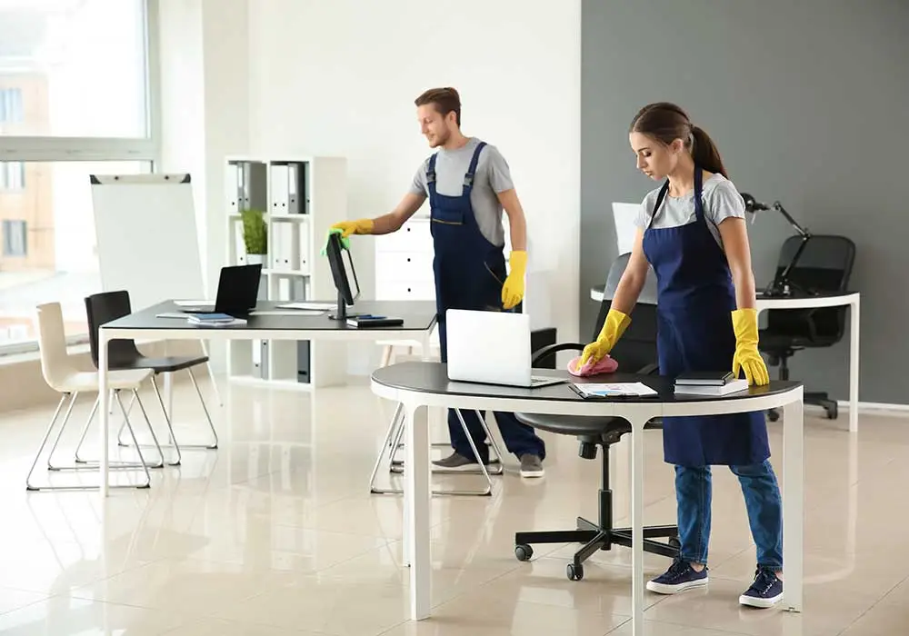 Impresa di pulizie Antonella Pulizia uffici. Due operatori con grembiule blu e guanti gialli intenti a pulire le scrivanie e i computer di un ufficio illuminato da ampie vetrate