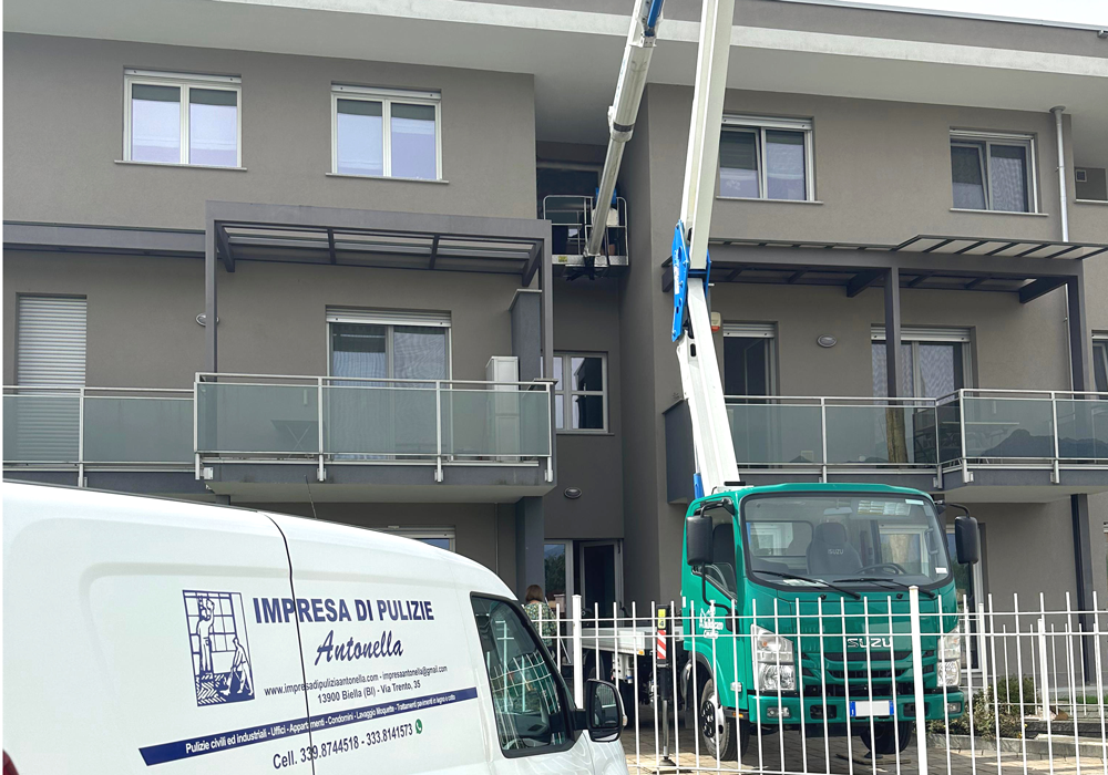 Impresa di pulizie Antonella Pulizia vetrate con l'ausilio di cestelli o piattaforme aeree. Furgone Bianco di Impresa di pulizie Antonella di fronte ad un condominio a due piani in cui un operatore dentro ad un cestello manovrato da un camion con braccio allungabile intento nella pulizia di una vetrata