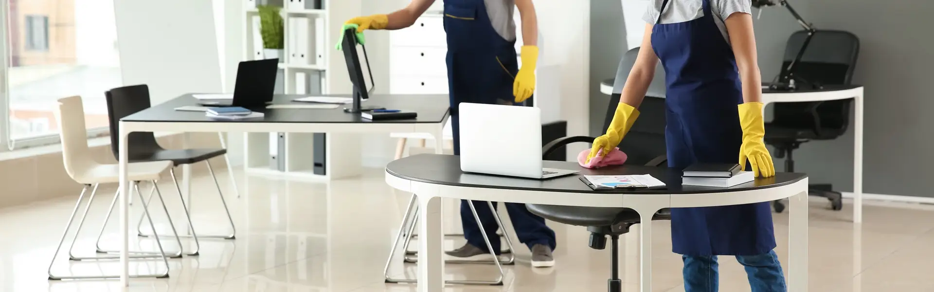 Impresa di pulizie Antonella Pulizia uffici. Due operatori con grembiule blu e guanti gialli intenti a pulire le scrivanie e i computer di un ufficio illuminato da ampie vetrate