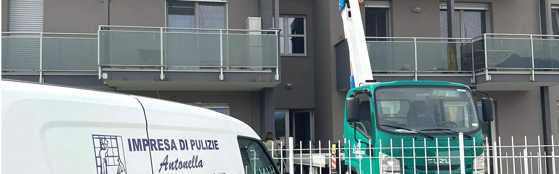 Impresa di pulizie Antonella Pulizia vetrate con l'ausilio di cestelli o piattaforme aeree. Furgone Bianco di Impresa di pulizie Antonella di fronte ad un condominio a due piani in cui un operatore dentro ad un cestello manovrato da un camion con braccio allungabile intento nella pulizia di una vetrata