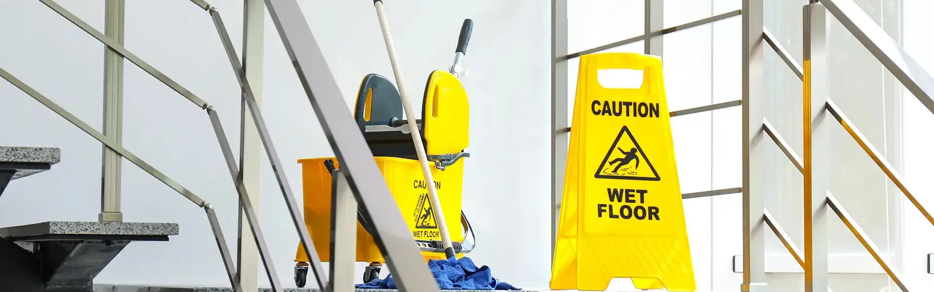Impresa di Pulizie Antonella Pulizia Condomini. Classico secchio delle pulizie giallo con rotelle con sopra appoggiato uno scopettone blu e cartello sempre giallo con su scritto caution wet floor fotografati sul pianerottolo di una rampa di scale